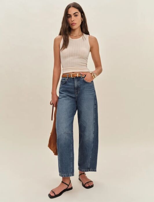 A woman stands against a plain background wearing a sleeveless beige top, high-waisted blue jeans, a brown belt, black sandals, and holding a brown bag.