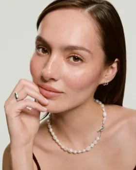 Woman with straight brown hair wears a pearl necklace, silver hoop earrings, and a silver ring, resting her hand on her chin and looking at the camera against a light background.