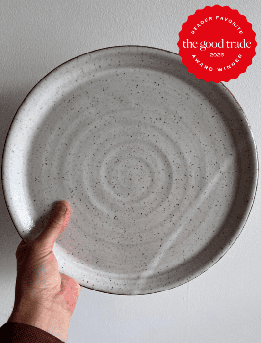 A hand holds a large, speckled ceramic plate against a white wall. A red badge in the corner reads "the good trade 2026 award winner, reader favorite.