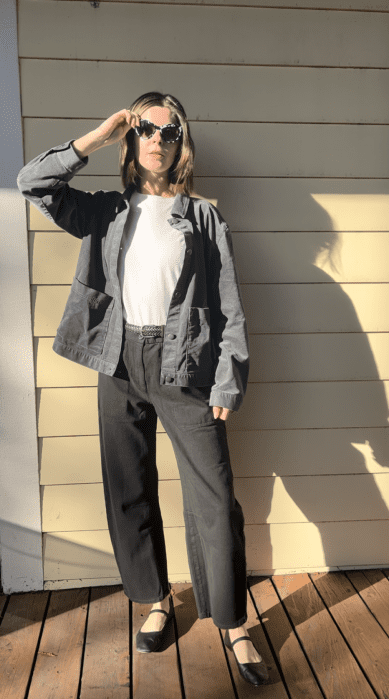 Person wearing sunglasses, a gray jacket, white t-shirt, black pants, and black shoes stands on a wooden deck in sunlight, casting a shadow on a light-colored wall.