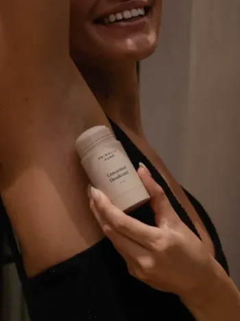 A woman applies Primally Pure Unscented Deodorant stick to her underarm while smiling.
