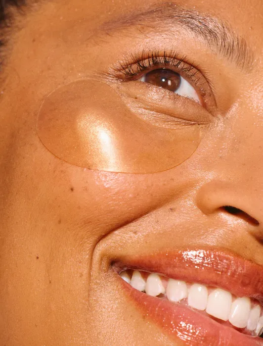 A close-up of a smiling person’s face, showing a transparent under-eye patch on their cheekbone and glossy lips.