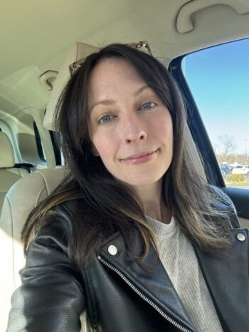 A woman with straight brown hair and a black leather jacket sits in a car, looking at the camera. Sunlight is coming through the window, and trees are visible outside.