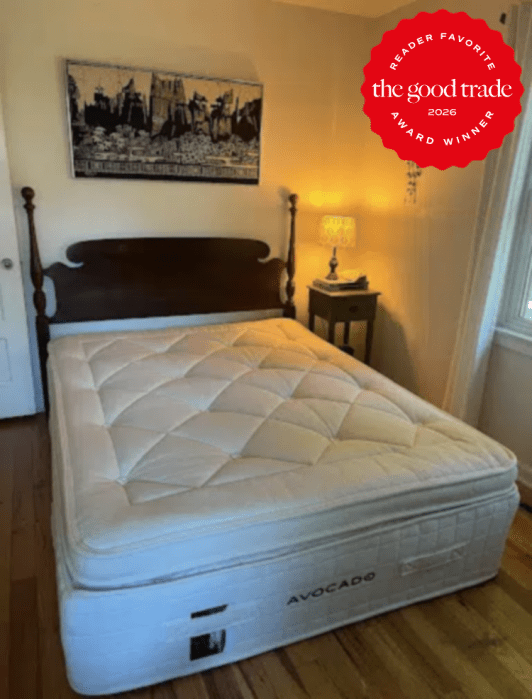 A neatly made bed with an Avocado mattress in a bedroom, beside a wooden nightstand with a lamp. A black-and-white cityscape artwork hangs above the headboard.