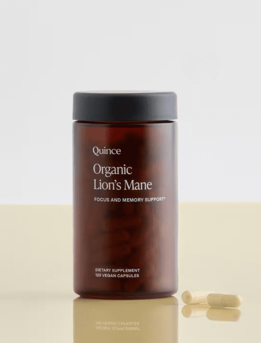 A brown jar labeled “Quince Organic Lion’s Mane” dietary supplement with two capsules lying beside it on a light surface.