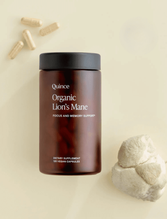 A brown bottle labeled “Quince Organic Lion’s Mane” sits on a pale surface with loose capsules and a piece of lion’s mane mushroom nearby.