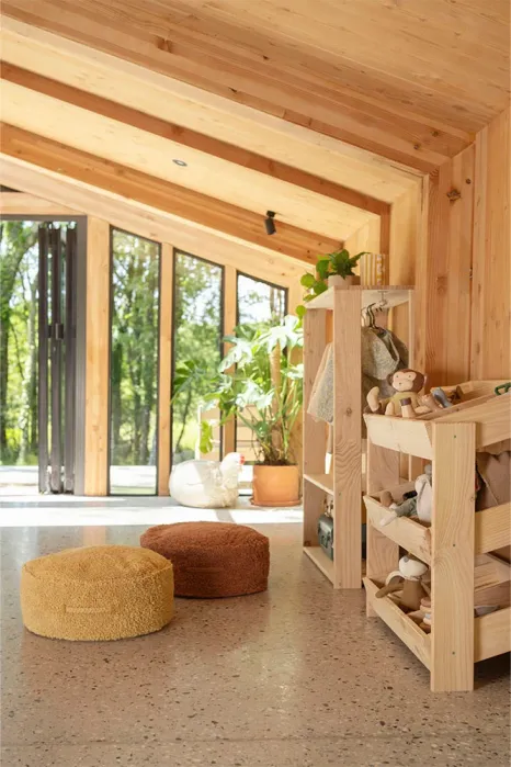 A wooden playroom with shelves holding toys, pouf cushions on the floor, and large windows overlooking green trees and plants outside.
