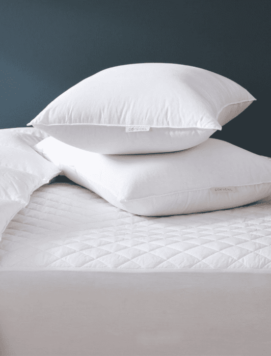 Two white pillows stacked on a neatly made bed with a white quilted mattress cover and a white comforter, against a dark blue wall.