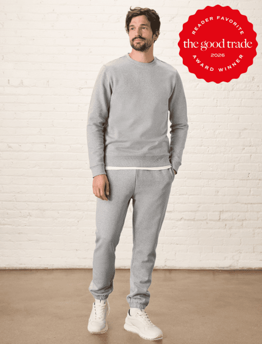 A man stands against a white brick wall wearing a grey sweatshirt, matching sweatpants, and white sneakers. A red badge in the corner reads "The Good Trade Award Winner 2026.