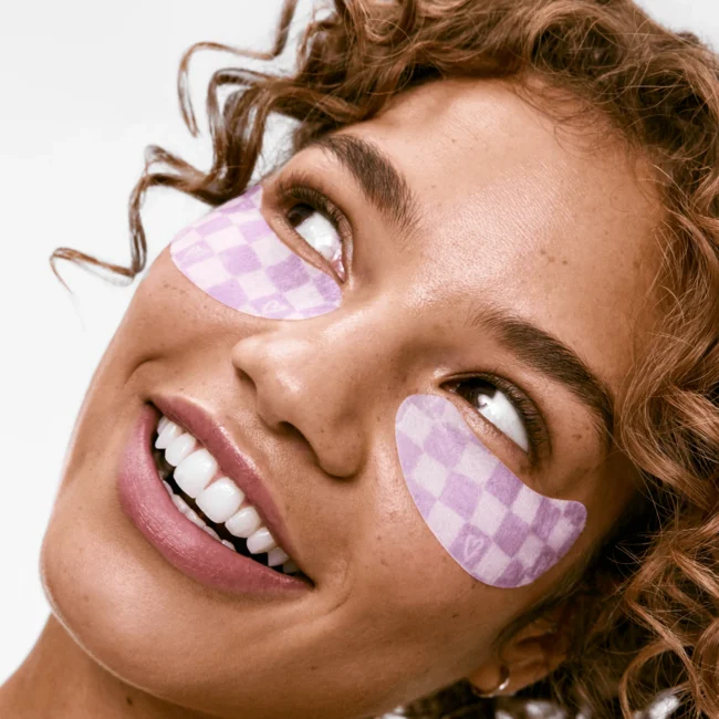Smiling person with curly hair wearing purple checkered under-eye patches, looking upwards.