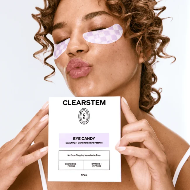 Woman with curly hair wears under-eye patches and holds a CLEARSTEM Eye Candy eye patch box close to her face, eyes closed, in a well-lit setting.
