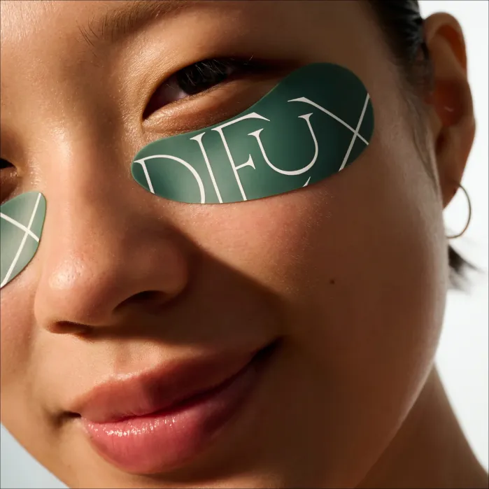 Close-up of a person wearing green under-eye patches with the word “DIEUX” printed on them. The person has clear skin, minimal makeup, and a small hoop earring.
