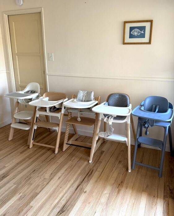 Four high chairs in different styles and colors are lined up against a wall on a wooden floor, with a framed fish print hanging above.