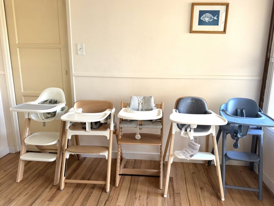Four modern high chairs with wooden legs and plastic seats in white, gray, and blue are lined up against a cream-colored wall in a room with wooden flooring.