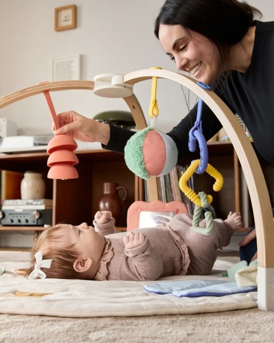 A woman smiles while playing with a baby lying on a play mat with hanging toys in a living room.