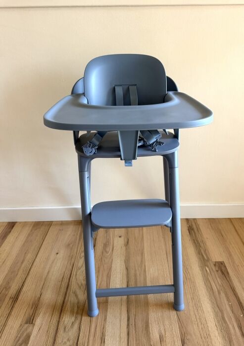 A modern gray high chair with tray and safety harness stands on a wooden floor against a plain light-colored wall.