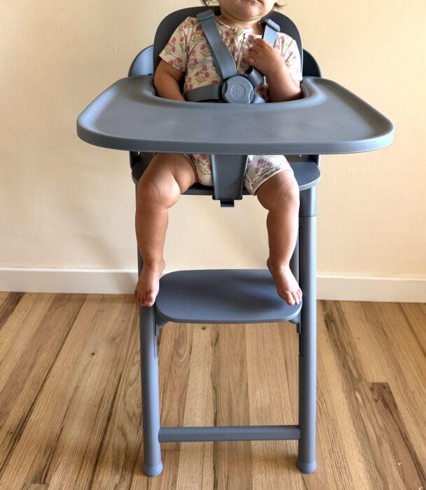 A baby wearing a floral onesie sits in a gray high chair with a safety harness, feet resting on the footrest, against a light wall and wooden floor.