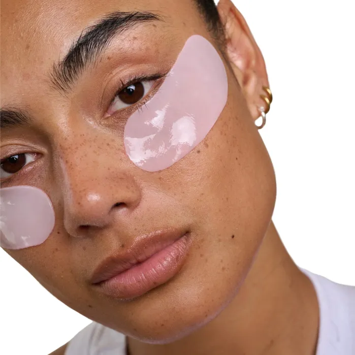 A person with under-eye gel patches looks at the camera. They have clear skin, natural makeup, and wear a gold hoop earring. The background is white.