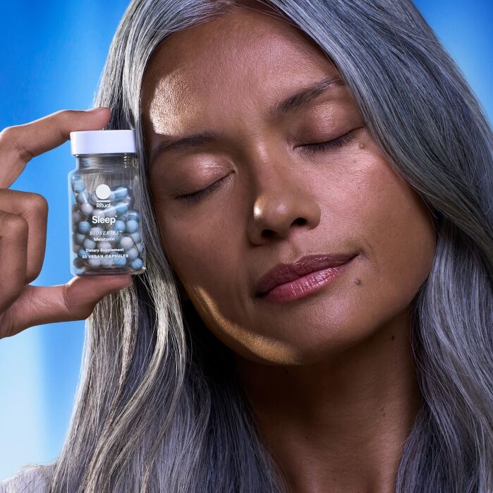 A woman with long gray hair and closed eyes holds a jar labeled "Sleep" with blue and white capsules against a blue background.