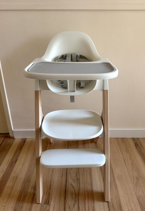 A white and light wood high chair with a gray tray is positioned on a wooden floor against a beige wall.