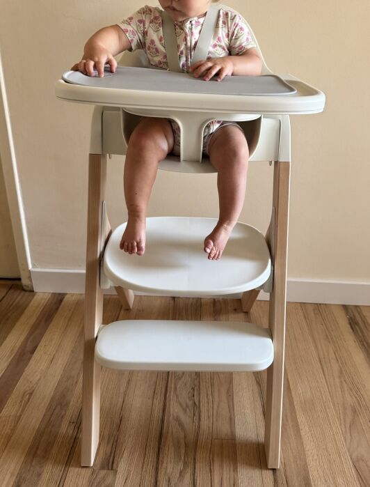 A baby sits in a modern high chair with a tray, wearing a patterned outfit. Only the baby’s torso, arms, and legs are visible. The chair is on a wooden floor against a plain wall.