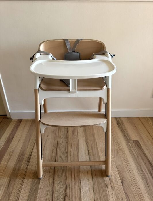 A wooden high chair with a white plastic tray and a gray safety harness is placed on a wooden floor against a light-colored wall.