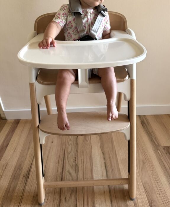 A young child sits in a wooden high chair with a white tray, wearing a floral-patterned shirt and a safety harness, feet resting on the footrest.