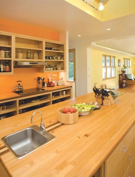 Spacious kitchen with a wooden island, sink, various utensils, and open shelves stocked with jars and cookware; adjoining open dining and living area with large windows.