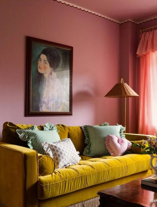 A living room with pink walls, a yellow velvet sofa with patterned cushions, a framed portrait, a wall sconce, a table lamp, pink curtains, and a vase of flowers on a table.