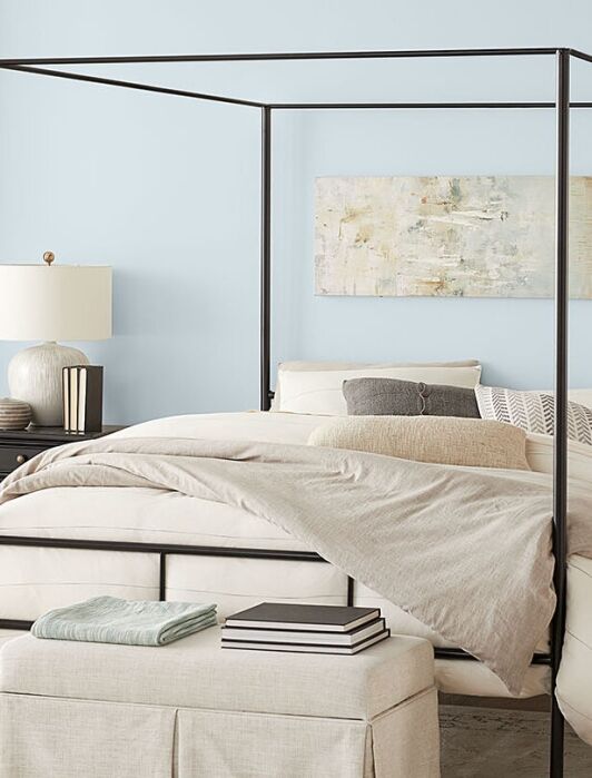 A modern bedroom with a black canopy bed, neutral bedding, two matching nightstands with lamps, and a bench at the foot of the bed, set against light blue walls.
