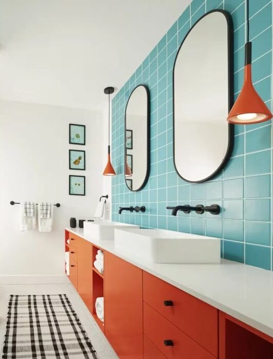 Modern bathroom with bright orange cabinets, double white sinks, oval mirrors, blue wall tiles, and orange pendant lights. Black fixtures and a black-and-white checkered rug are present.