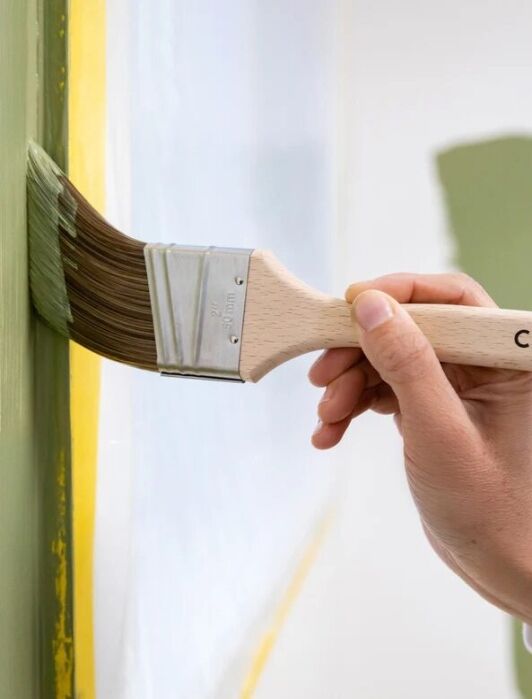 A hand holds a paintbrush labeled "CLARE," painting a green stripe along the edge of a wall with painter’s tape applied at the border.