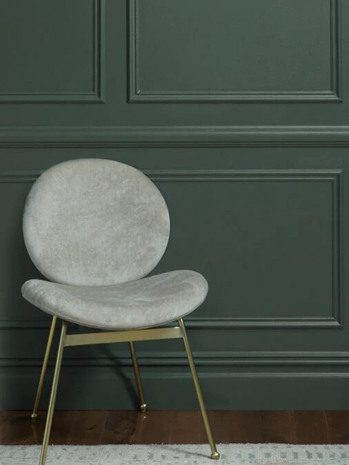A modern light gray upholstered chair with gold legs sits on a rug in front of a dark green paneled wall.
