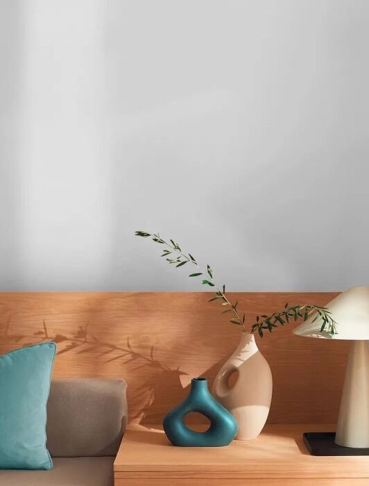 A wooden side table with a beige lamp, two decorative vases with green branches, and a blue cushion on a beige sofa, set against a plain light gray wall.