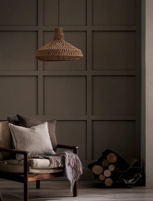 A cozy armchair with cushions and a throw sits beside a small table and a stack of firewood under a woven pendant light in a neutral-toned room with paneled walls.