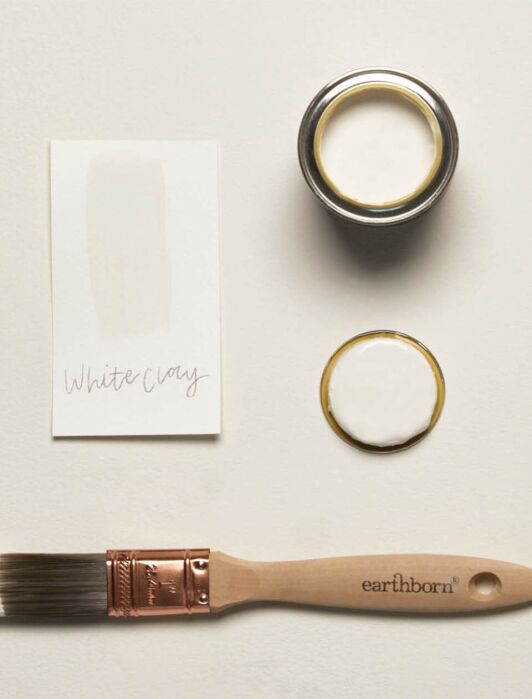 A paintbrush, a paint sample labeled "White Clay," an open paint can with white paint, and a lid are arranged on a light surface.