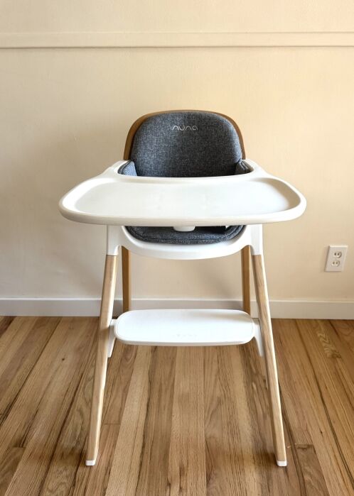 A modern high chair with a white tray and wooden legs stands on a hardwood floor against a beige wall with an electrical outlet visible on the right.