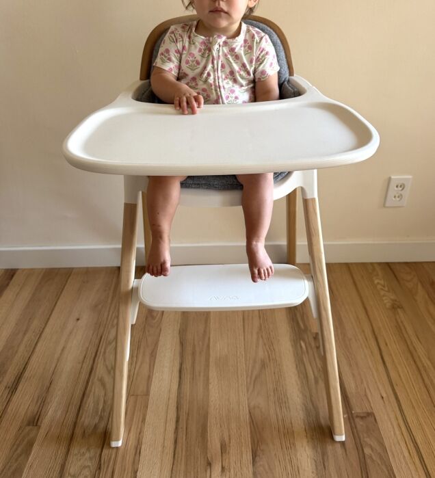 A young child sits in a high chair with a white tray and wooden legs on a wooden floor, facing the camera.
