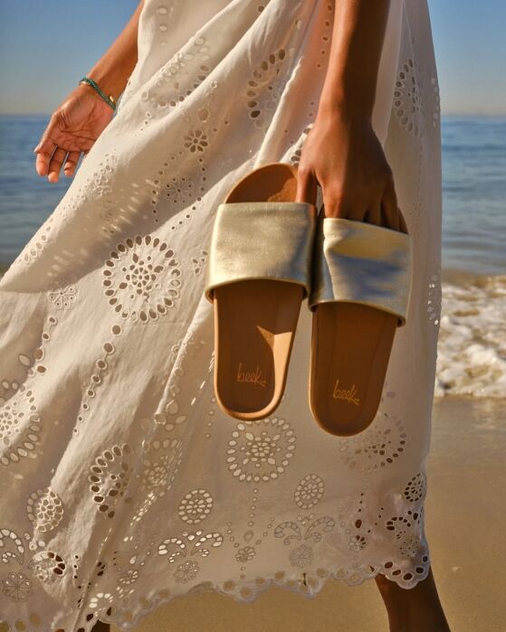 A person in a white eyelet dress stands by the shore, holding a pair of gold slide sandals in one hand.