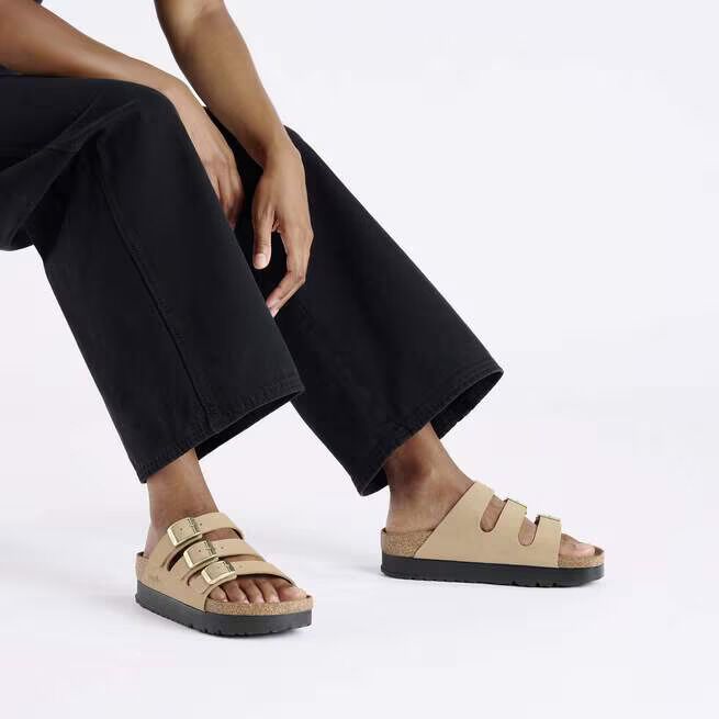 A person wearing black wide-leg pants and beige three-strap sandals sits with one hand resting on their knee against a plain white background.