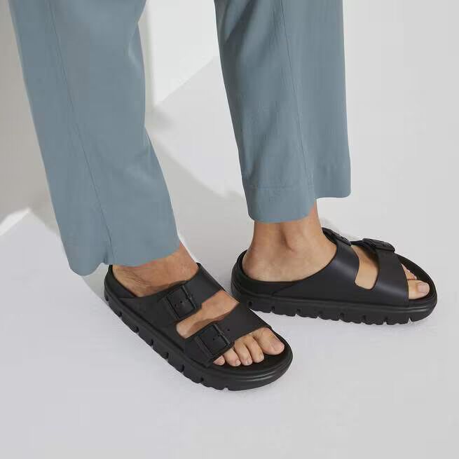 Person wearing light blue pants and black slide sandals with two buckle straps, standing on a white surface.