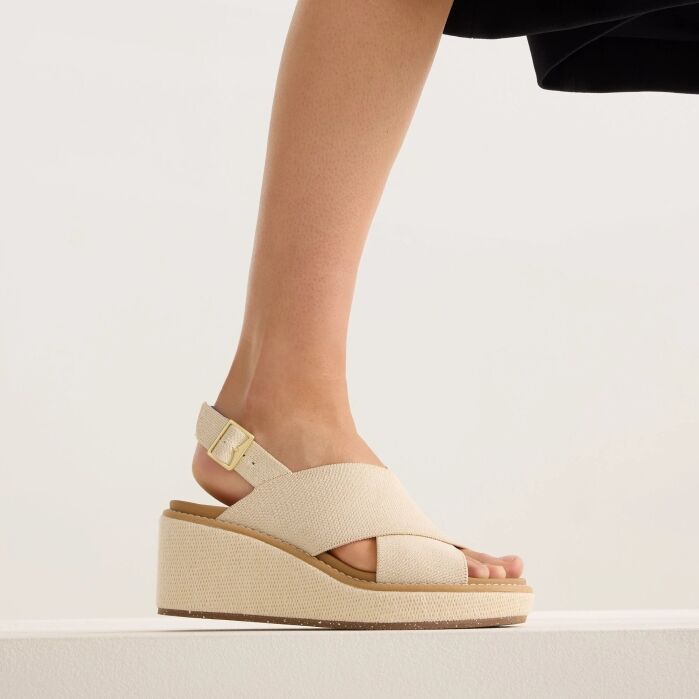 A person wearing beige, open-toe, platform wedge sandals with a gold buckle strap stands against a neutral background.