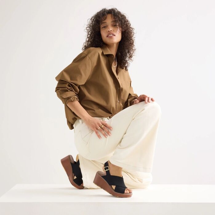 Woman with curly hair poses on a white surface, wearing a brown button-up shirt, white wide-leg pants, and black wedge sandals.