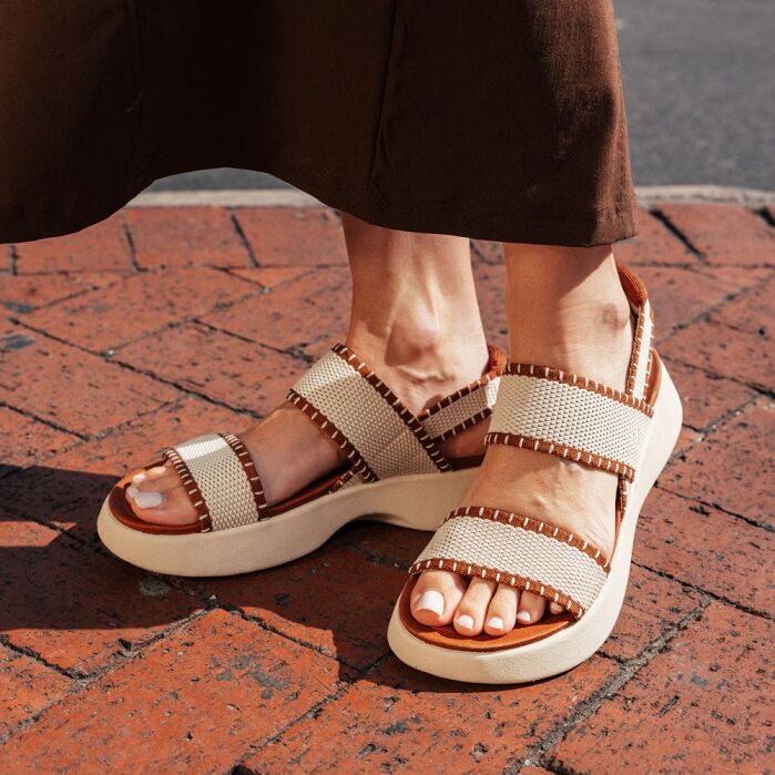 Person wearing beige and brown woven sandals with thick soles, standing on a red brick pavement, part of a brown skirt visible.