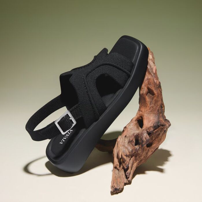 A black sandal with a fabric strap and buckle is displayed balanced on a piece of driftwood against a gradient beige background.