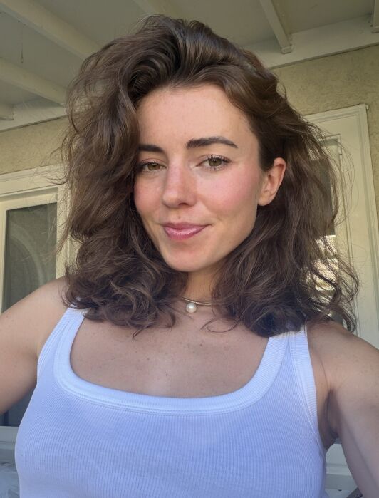 Woman with wavy brown hair wearing a white tank top and a pearl necklace, standing indoors near a window and smiling slightly at the camera.