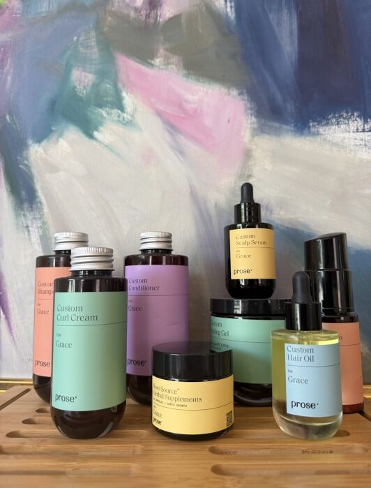 Various custom-labeled Prose hair and skin products, including bottles, jars, and dropper vials, are arranged on a wooden tray against an abstract colorful background.