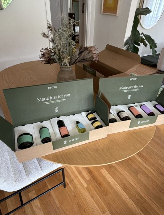 Two open Prose boxes with personalized hair care products sit on a round wooden table next to a dried flower arrangement in a vase.