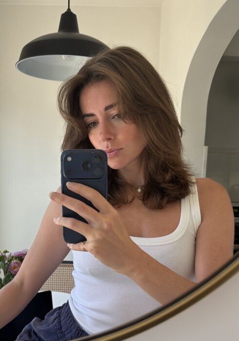 Woman with shoulder-length brown hair, wearing a white tank top, takes a mirror selfie with a smartphone in a well-lit room. A pendant light and some flowers are visible in the background.