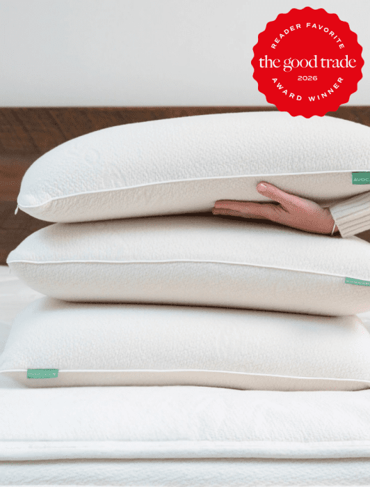 A person holding a stack of three white pillows on a bed with a red badge in the corner reading "the good trade award winner 2026.